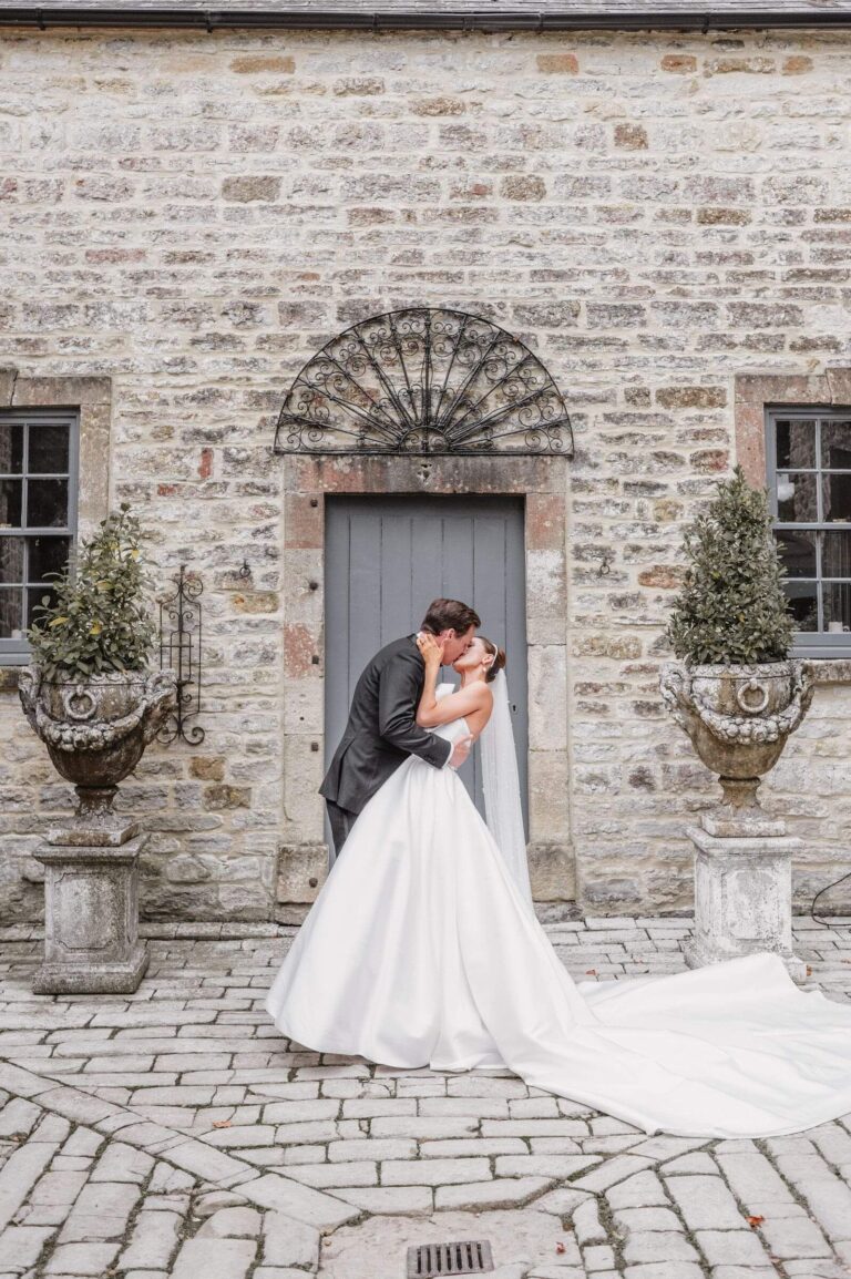 A bride and groom kissing in a cobbled stone courtyard, her train spread behind, Kate Edmondson Bridal Couture