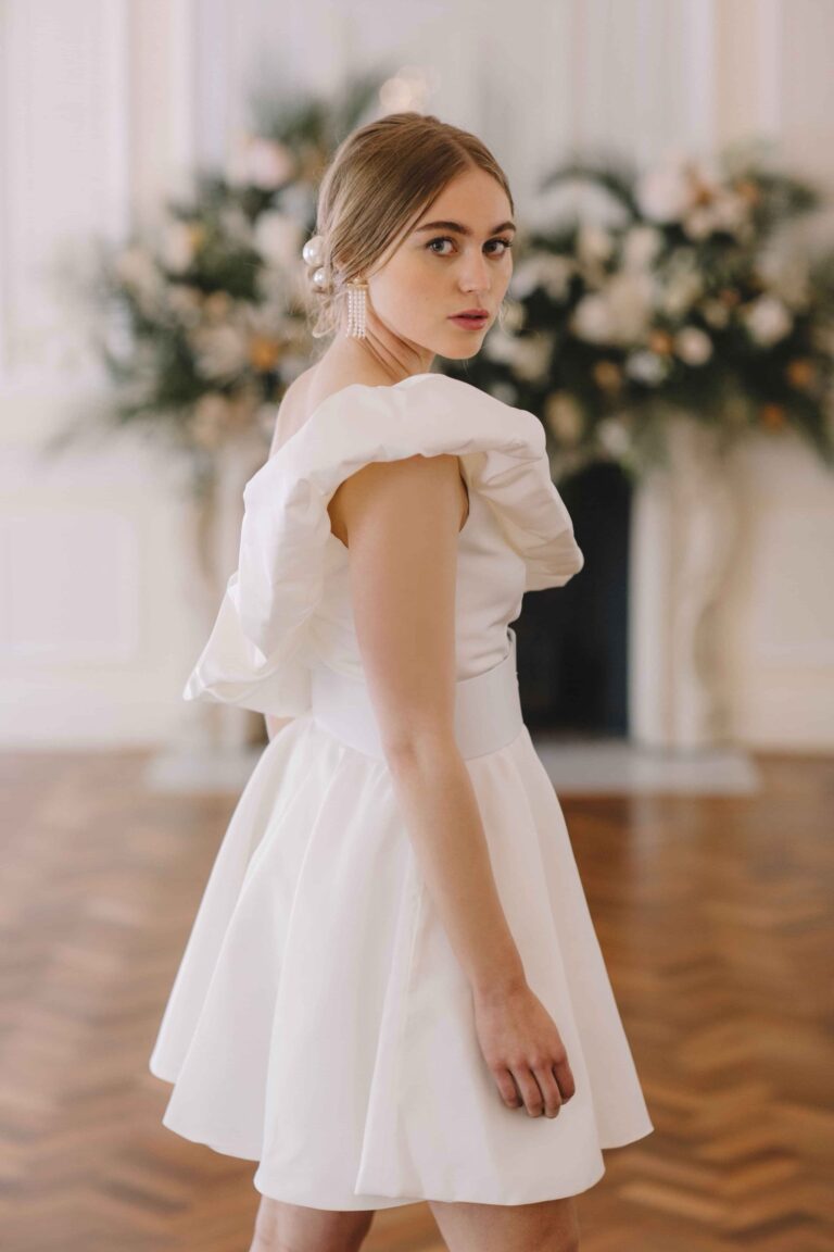 A short dress with a ruffled shoulder in a room of white flowers, Kate Edmondson Bridal Couture