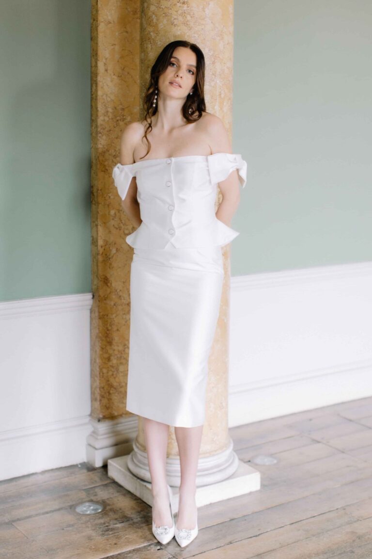A fitted off shoulder dress with peplum detail against pale green panelling, Kate Edmondson Bridal Couture