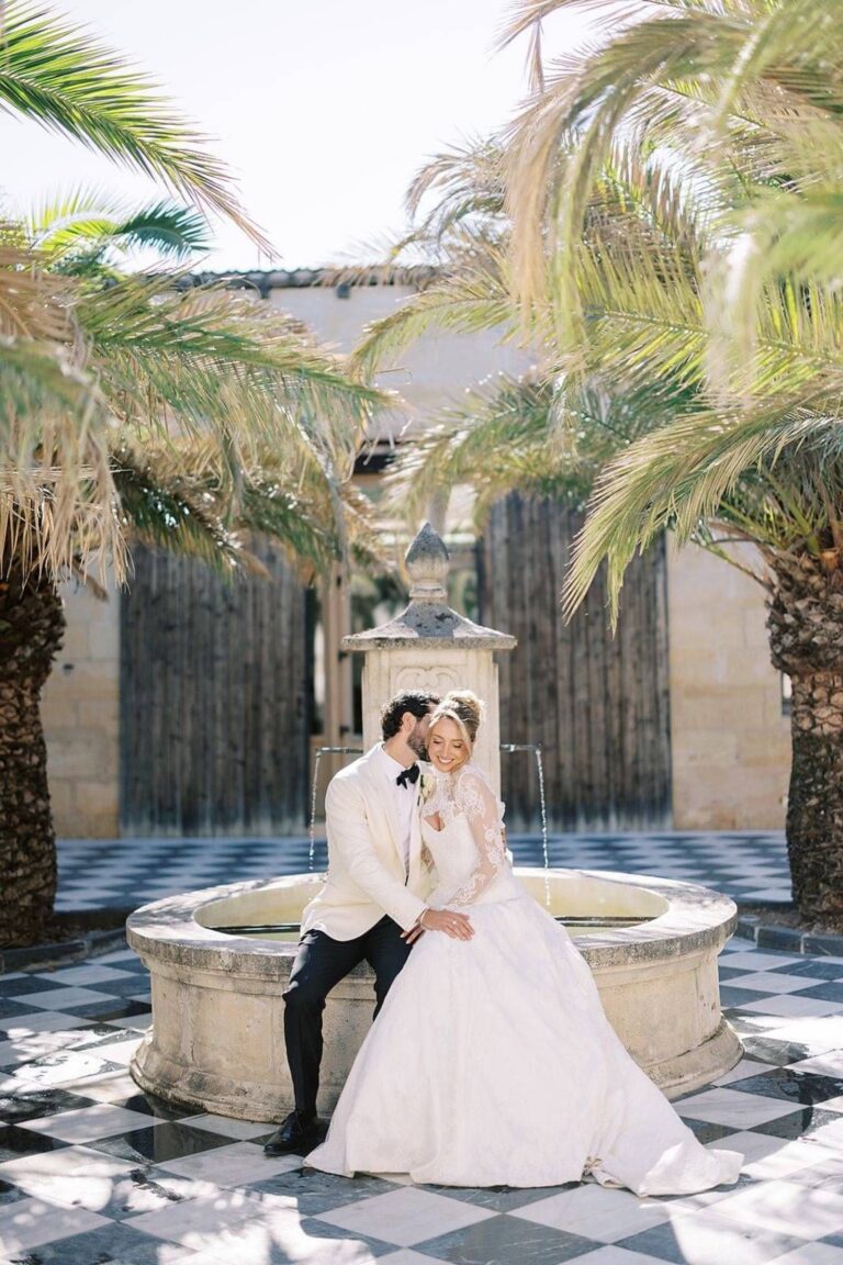 A bride and groom seated by a courtyard fountain among palm trees, Kate Edmondson Bridal Couture