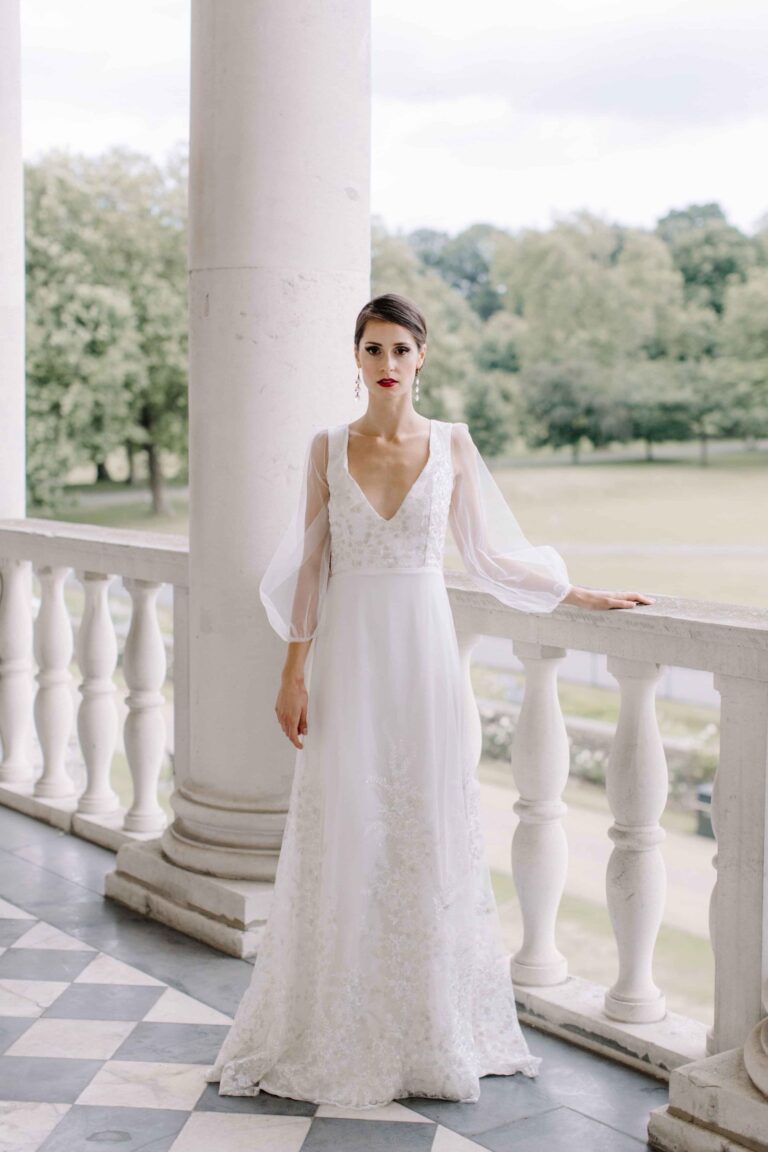 A V neck gown with sheer sleeves on a balustraded terrace, Kate Edmondson Bridal Couture