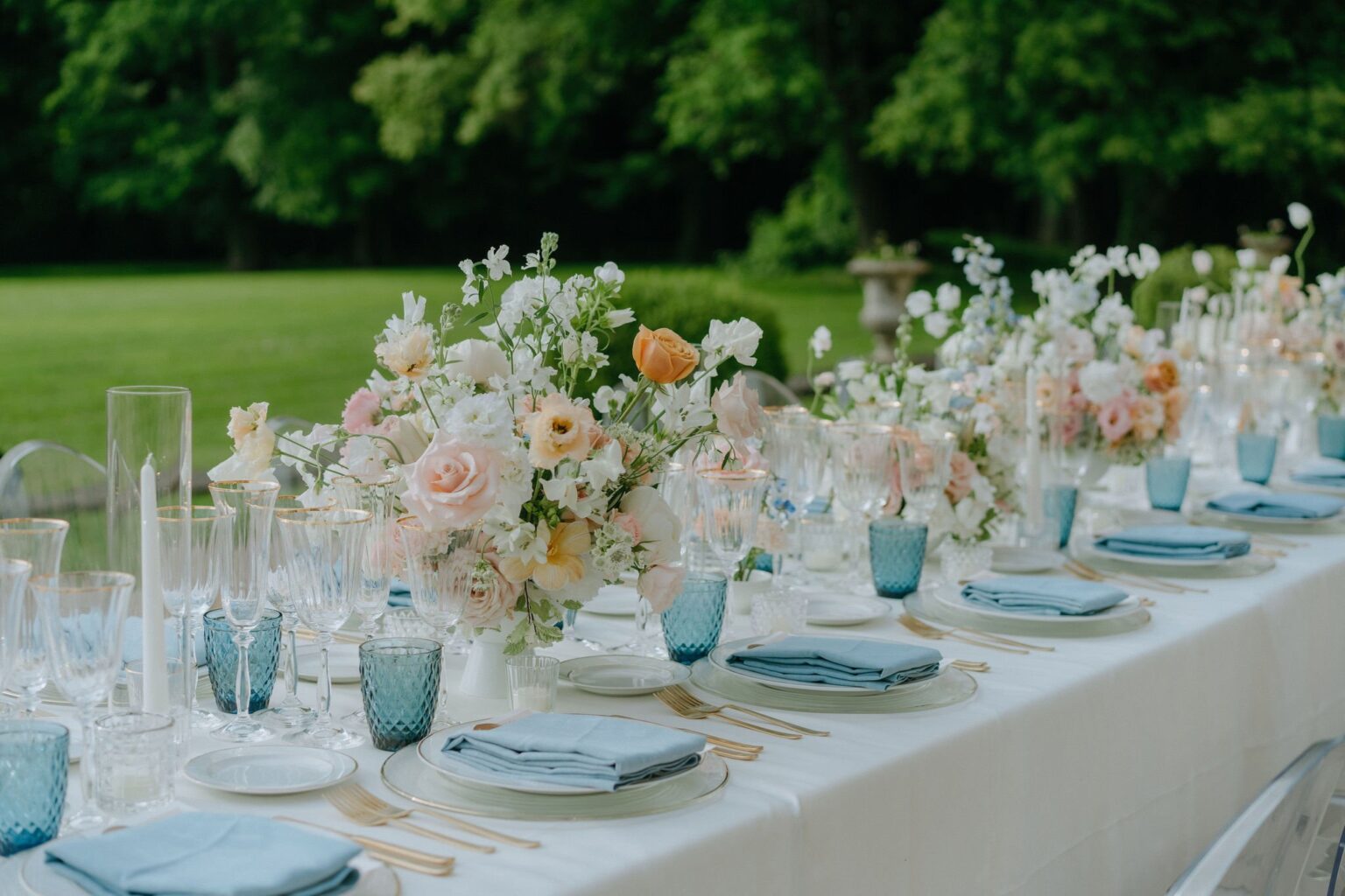 A long lawn table with blue glassware, blush flowers and white linen