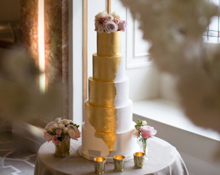 A tiered white wedding cake with gold leaf and pink roses, set by a window, Kimpton Fitzroy London
