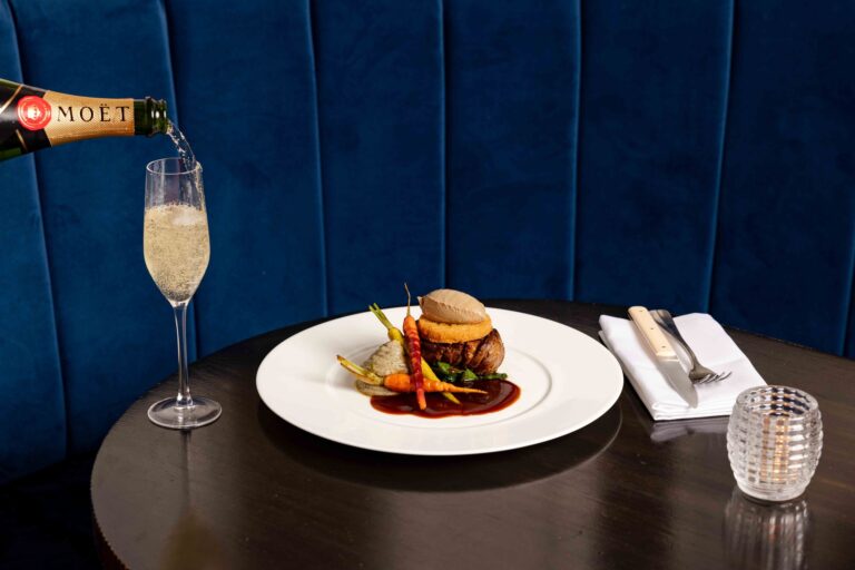 A plated main course with a glass of Champagne being poured at a blue velvet banquette, Kimpton Fitzroy London