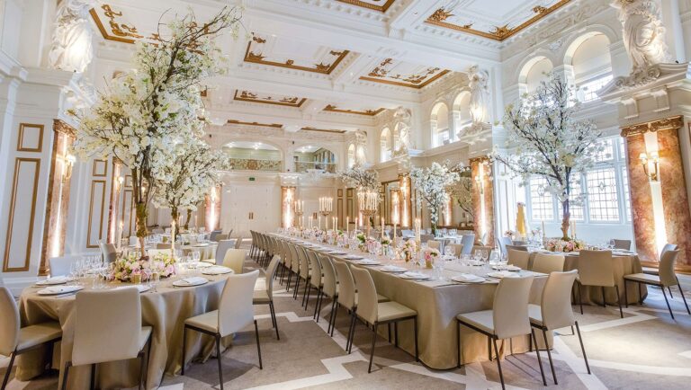 Long tables in the Ballroom dressed with neutral linen under tall blossom trees, with the gallery arches above, Kimpton Fitzroy London