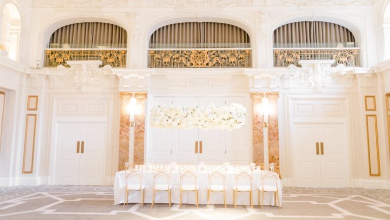 A small top table dressed in white with gold chairs and a white flower garland, set against the Ballroom's arched windows, Kimpton Fitzroy London