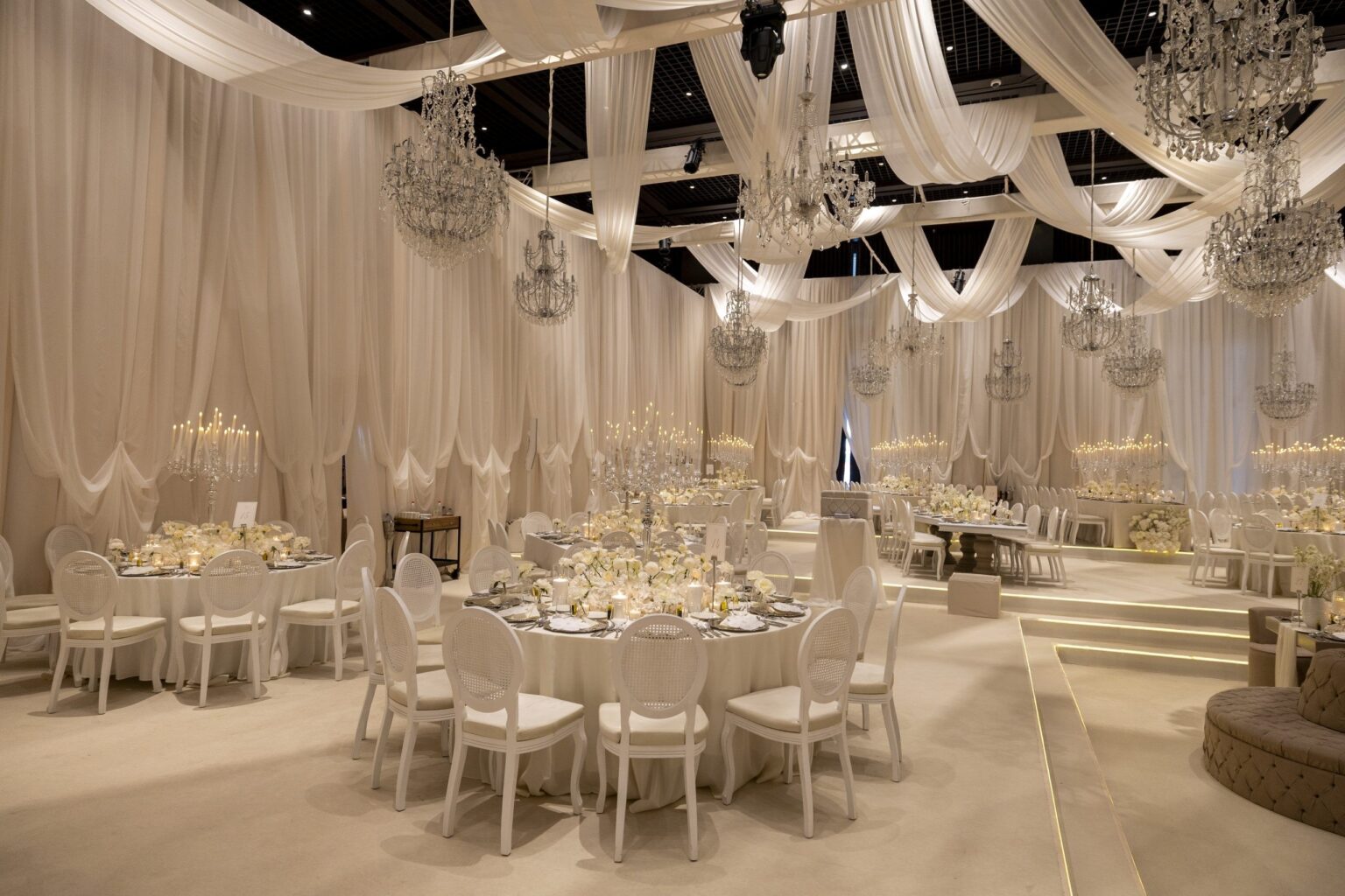 A white draped reception hall with chandeliers and round tables, KM Events
