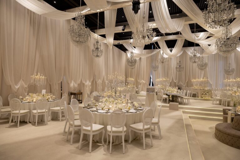 A white draped reception hall with chandeliers and round tables, KM Events