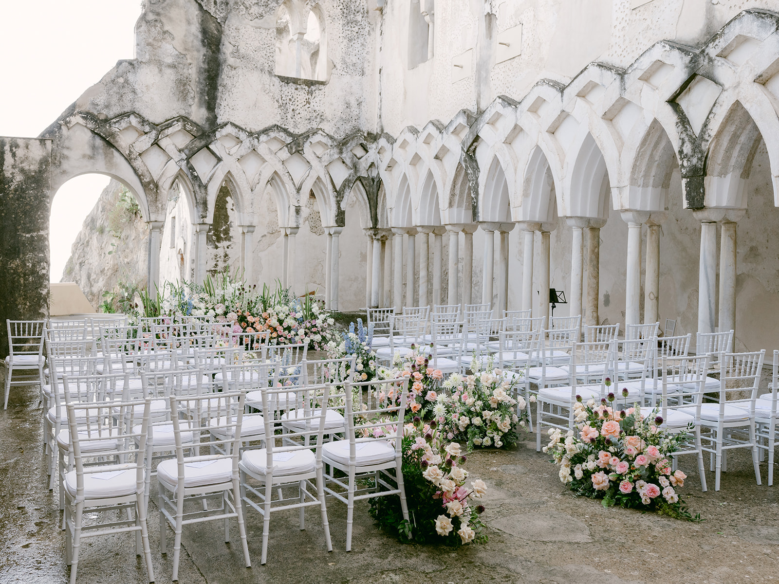 White chairs and pastel flower banks set for a ceremony in the cloister of Anantara Convento di Amalfi, La Calla Events
