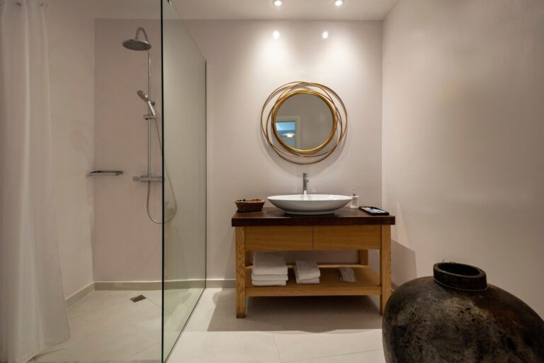 An ensuite with a glass walk-in shower, a gilt round mirror and a hand-thrown urn, La Maltese Estate