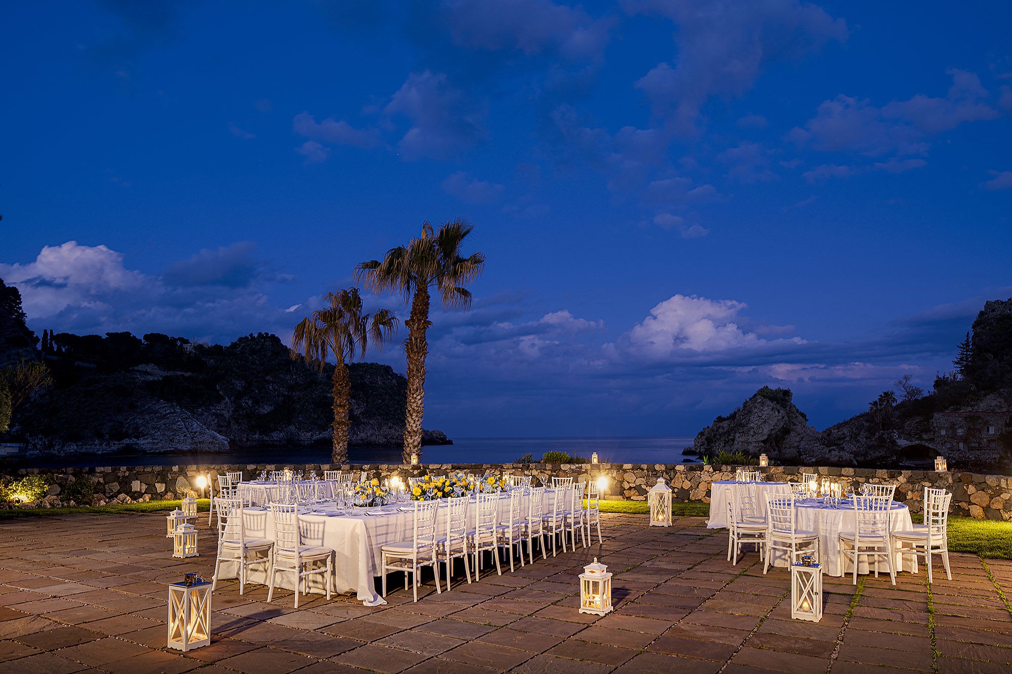 A long wedding table at blue hour, lanterns lit and the islet of Isola Bella behind, La Plage Resort