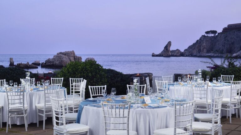 An evening reception on the terrace at dusk, pale blue linen and the rocks of the bay beyond, La Plage Resort