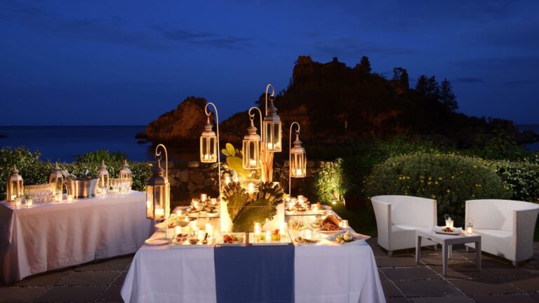 A candlelit beach dinner after dark, lanterns and the Isola Bella cliffs behind, La Plage Resort