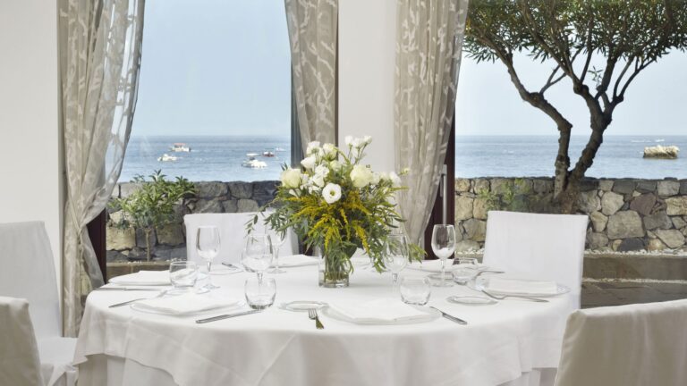 A dressed table at Idda, the sea framed through the window and an olive tree outside, La Plage Resort