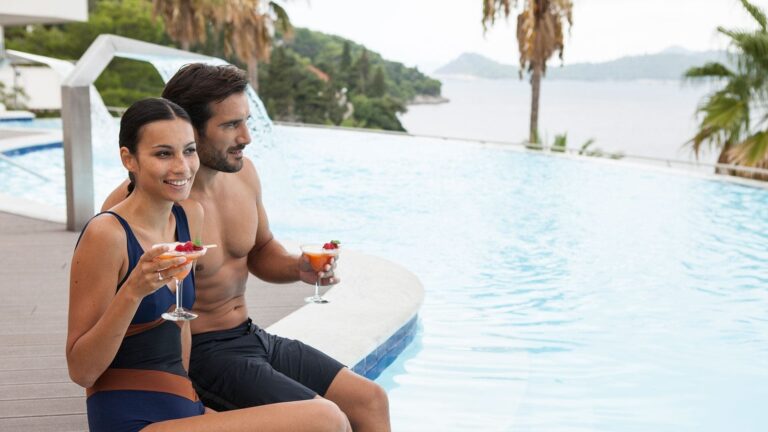 A couple at the pool edge with cocktails, the sea and the far shore beyond the water, Lafodia Sea Resort