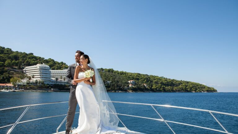 A bride and groom on the prow of a boat, the island and the resort behind them on the Adriatic, Lafodia Sea Resort