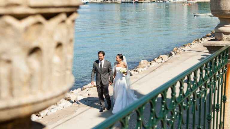 A couple walking a stone seafront balustrade, the bride in a pale gown beside the water, Lafodia Sea Resort