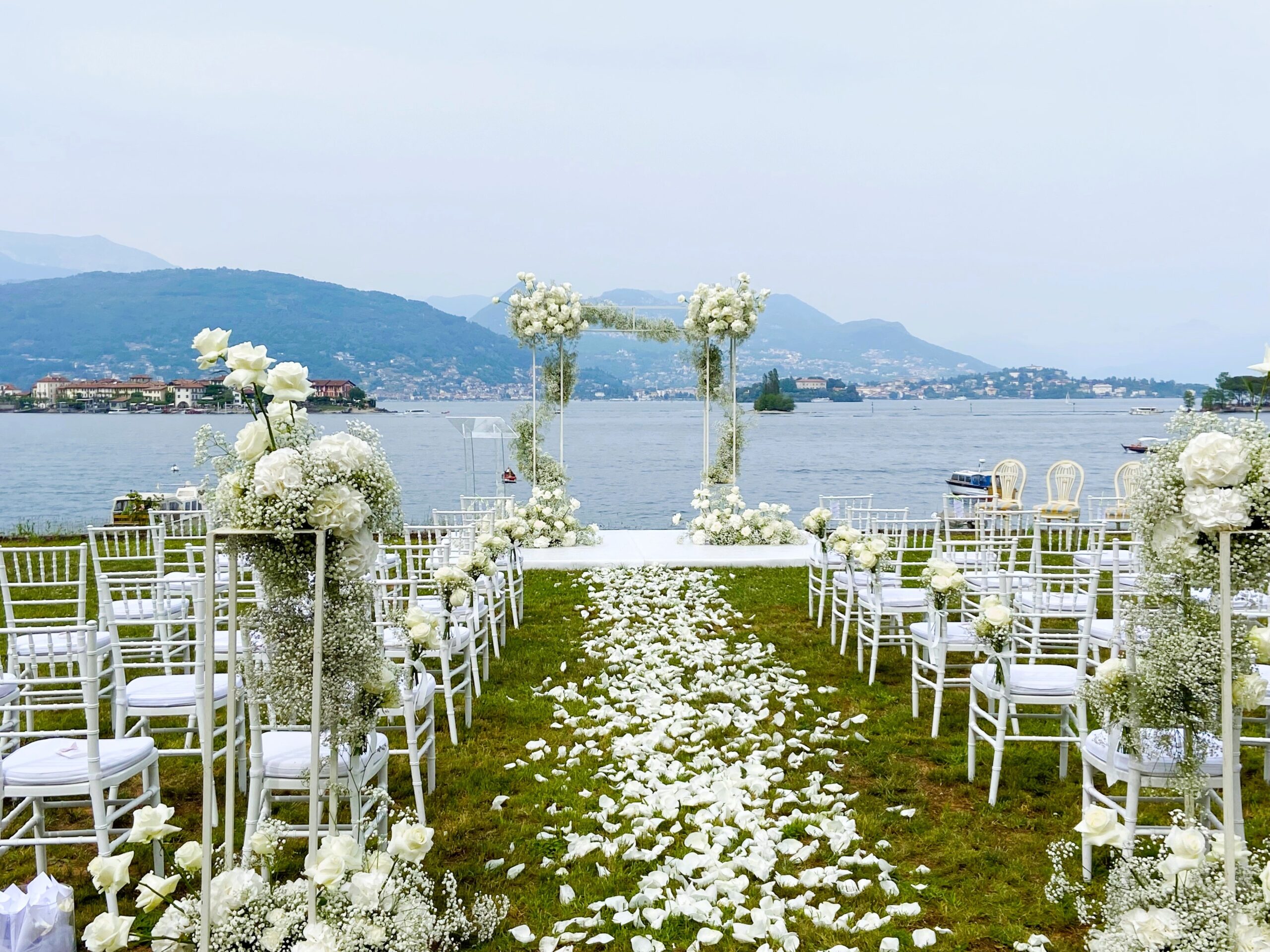 An aisle of white rose petals between columns of gypsophila leading to a slim floral arch, with the lake and an island beyond, Lake Maggiore, Lake Maggiore & Tuscany Weddings
