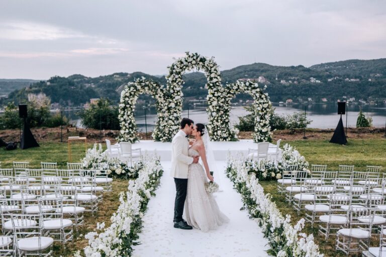 A white double floral arch at the head of a petal aisle on a lawn above the lake, the couple kissing beneath it, Lake Maggiore, Lake Maggiore & Tuscany Weddings