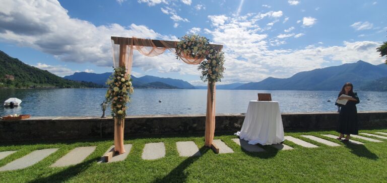 A timber arch dressed with white flowers and peach gauze at the water's edge, the registrar's table beside it, Lake Maggiore, Lake Maggiore & Tuscany Weddings