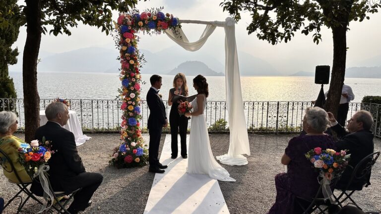 A civil ceremony on a lakeside terrace at sunset under an arch of blue and coral flowers with a white drape, Lake Maggiore, Lake Maggiore & Tuscany Weddings