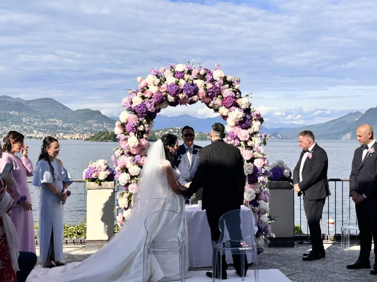 A ceremony on a lakeside terrace beneath a full arch of lilac and white hydrangea, guests standing, the far shore behind, Lake Maggiore, Lake Maggiore & Tuscany Weddings