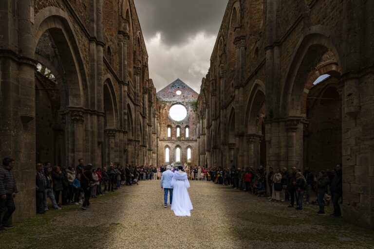 The couple walking away down the roofless nave of San Galgano Abbey between standing guests, Tuscany, Lake Maggiore & Tuscany Weddings