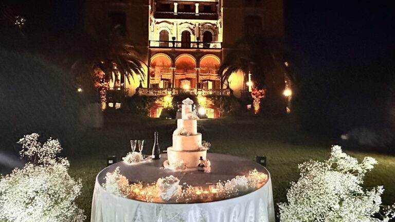 A tiered wedding cake lit at night on a round table banked with gypsophila, an illuminated villa facade behind, Lake Maggiore & Tuscany Weddings