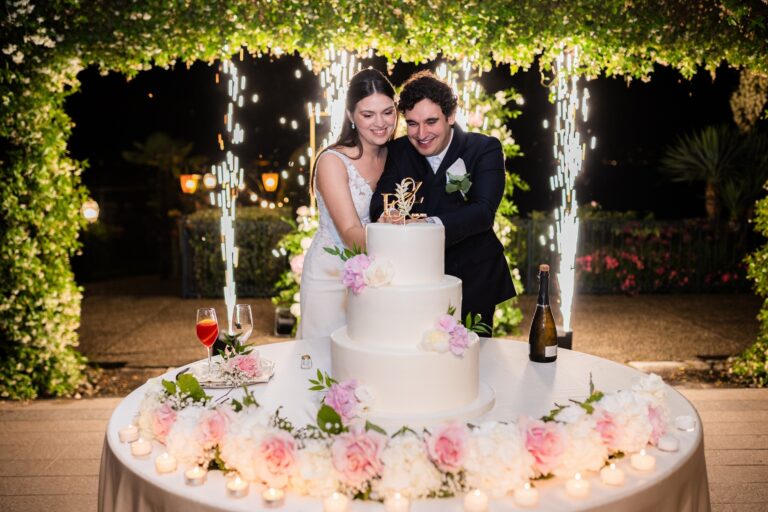 The couple cutting a white cake trimmed with pink roses under a greenery canopy between sparkler fountains, Lake Maggiore & Tuscany Weddings