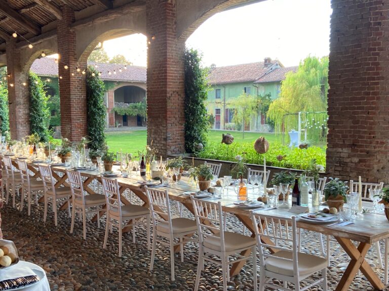 A long wooden dinner table under a brick portico, potted herbs down the centre, the farmhouse garden and willows beyond, Lake Maggiore & Tuscany Weddings