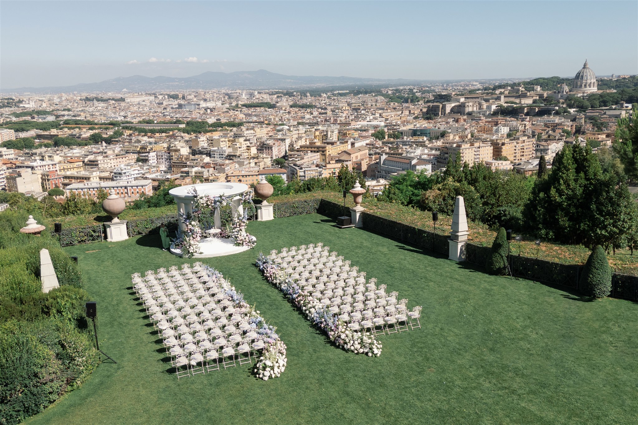 A ceremony laid out on a terraced lawn above Rome, white chairs in two blocks facing a flower-hung gazebo, with the dome of St Peter's on the skyline, Le Rêve