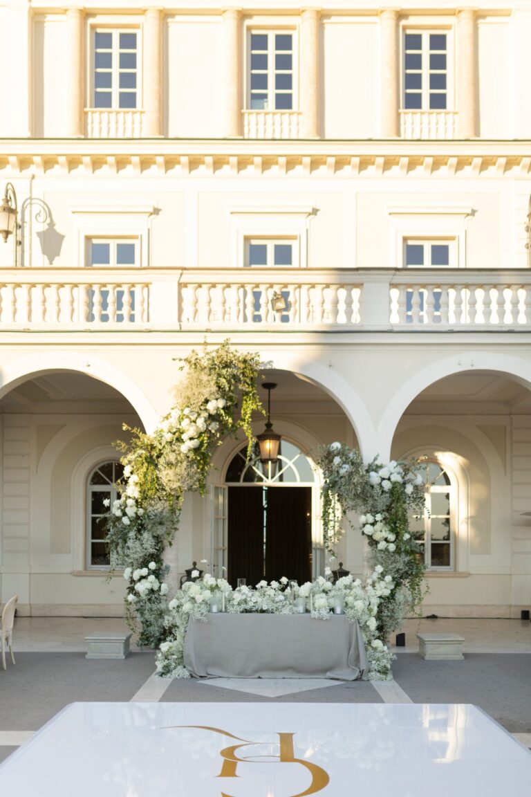 A ceremony set under the portico at Villa Miani, Rome, an arch of white flowers and greenery over the altar and a monogrammed floor, Le Rêve