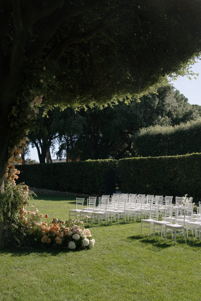 White ceremony chairs on a lawn beside clipped hedges, a tree wrapped in peach and white flowers at the front, Le Rêve