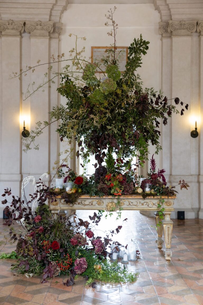A tall arrangement of dark foliage and deep red flowers over a gilded console in a palazzo hall, Le Rêve