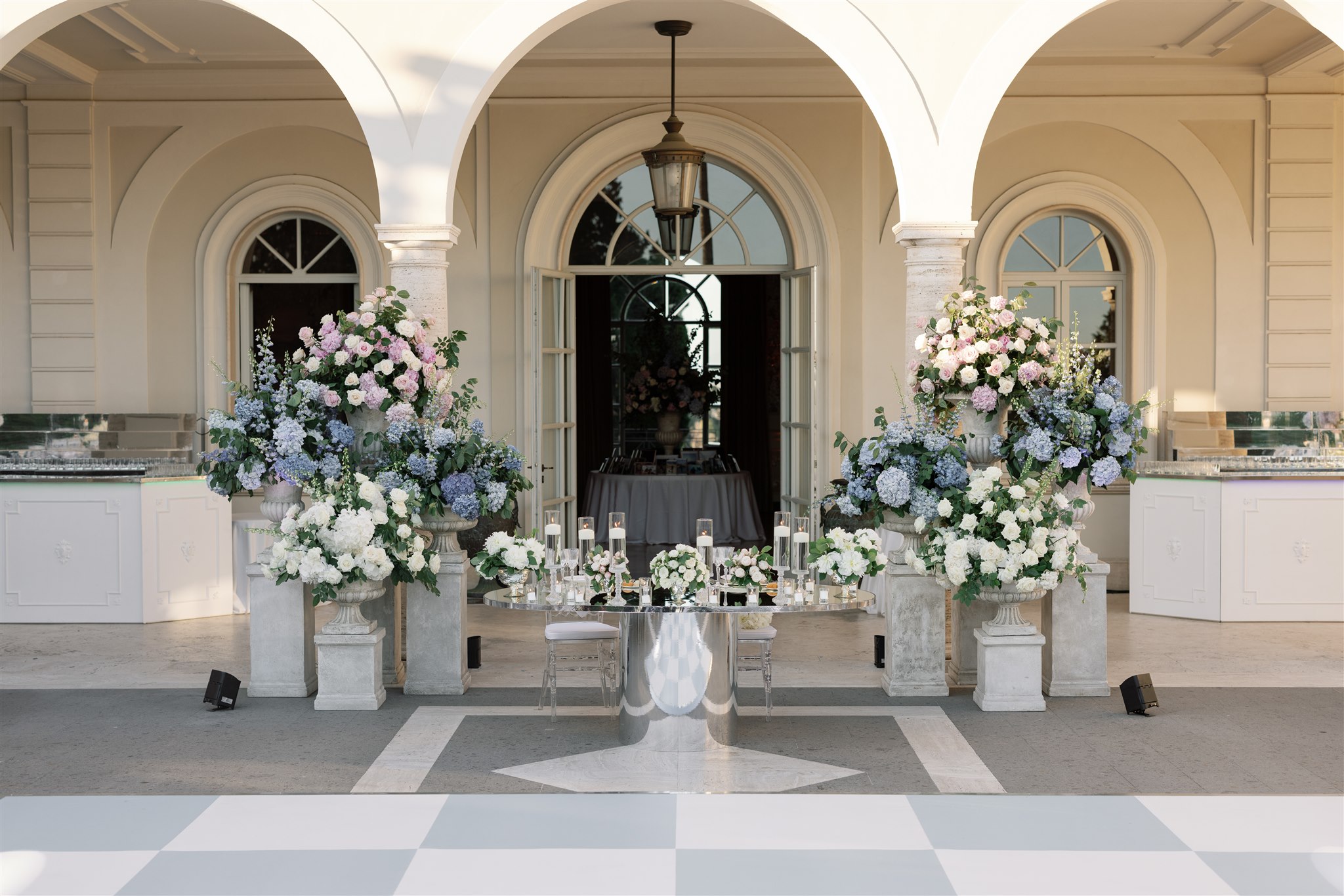 A table set under a villa portico, framed by tall urns of blue hydrangea and white roses, a chequerboard floor in front, Le Rêve