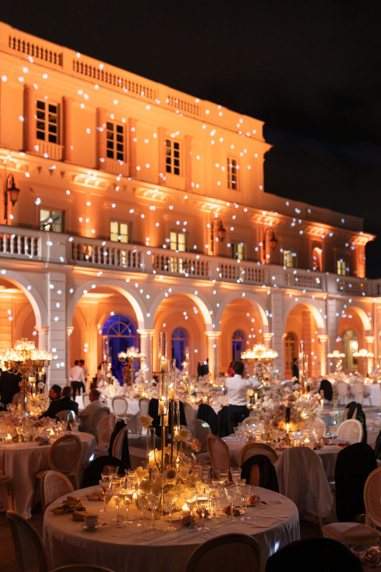 A villa facade lit gold after dark, round tables with candelabra on the terrace below, Le Rêve