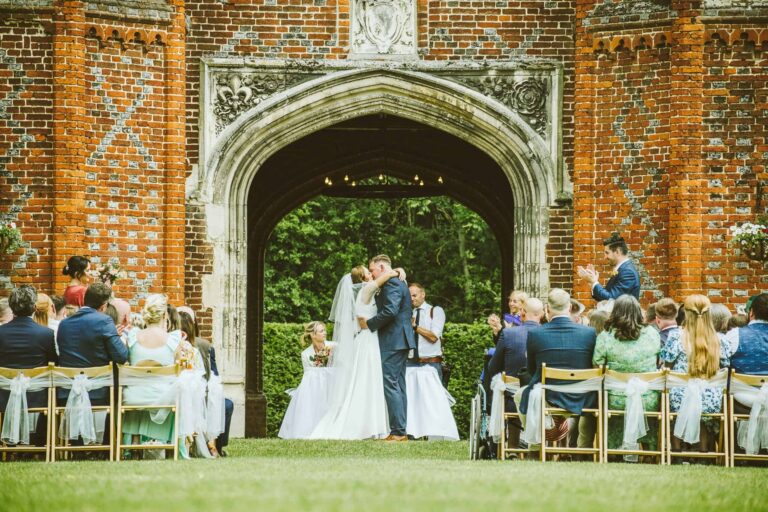 An outdoor ceremony on the lawn beneath the Great Tower, the couple sharing their first kiss before seated guests, Leez Priory