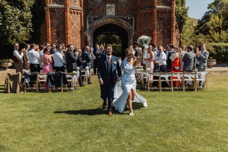 The newly married couple walking back down the aisle outdoors as guests cheer, the Tower behind, Leez Priory