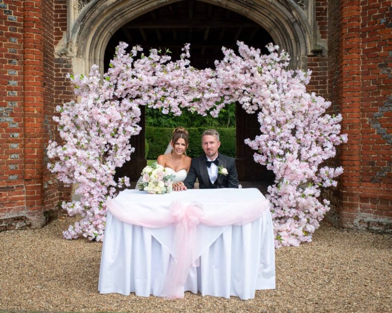 The couple at the signing table framed by a pink blossom arch within the Tower carriageway, Leez Priory