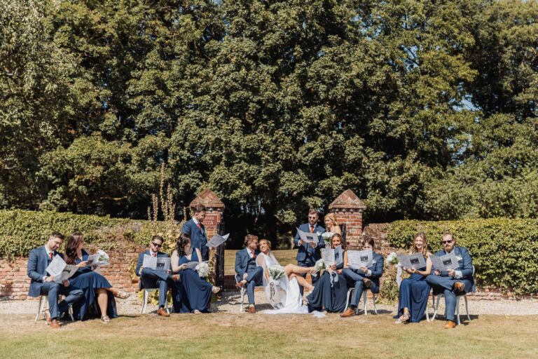 The whole wedding party seated in the garden, reading newspapers together in a playful group portrait, Leez Priory