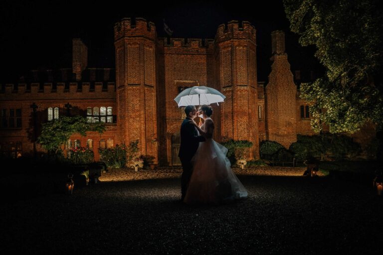A night portrait of the couple under a white umbrella before the floodlit Tudor mansion, Leez Priory