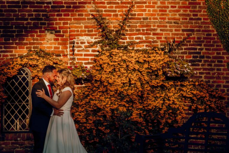 An evening portrait of the couple against a brick wall clad in autumn-orange pyracantha, Leez Priory