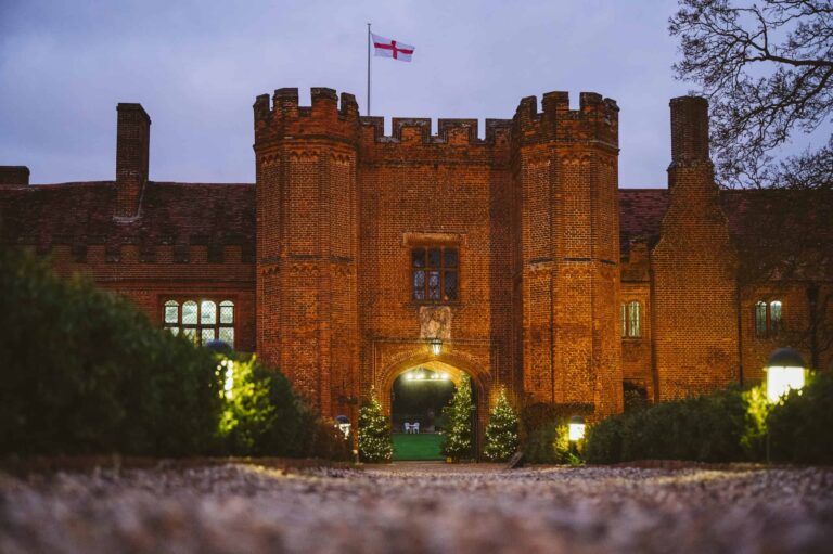 The Main House and its castellated towers floodlit at dusk, the England flag flying and the carriageway lit within, Leez Priory