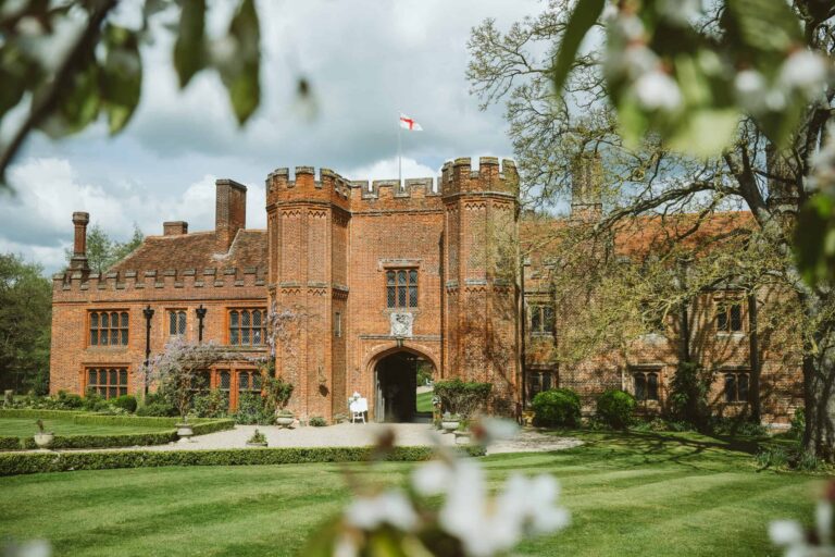 The house front by day, its towers and battlements framed by spring blossom, the England flag above, Leez Priory