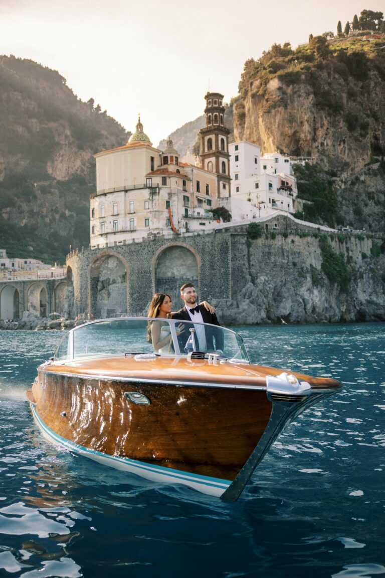 In a wooden launch on flat blue water, a cliff town stacked up the ravine behind, Leo Eventi