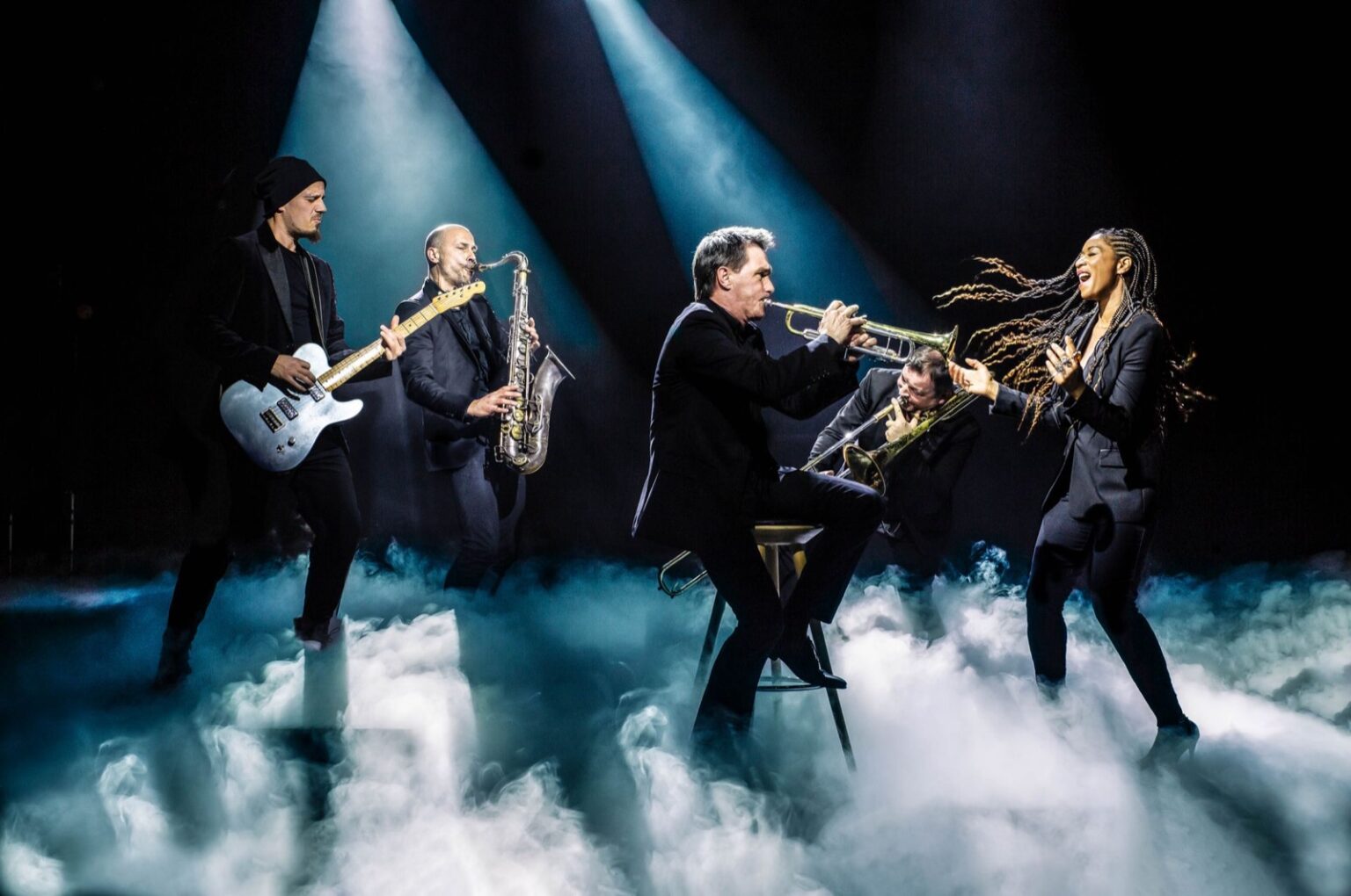 Five musicians on a smoky stage with saxophone, guitar and trumpet under cool spotlights