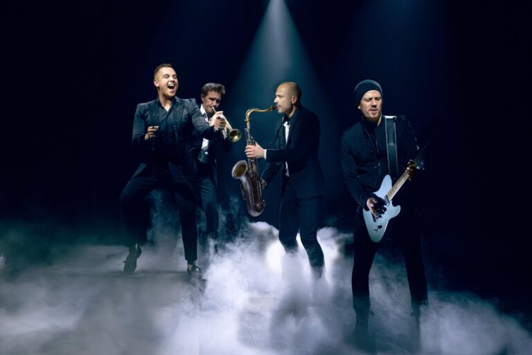Four band members in dark suits with saxophone and guitar on a hazy lit stage, Les Cigales