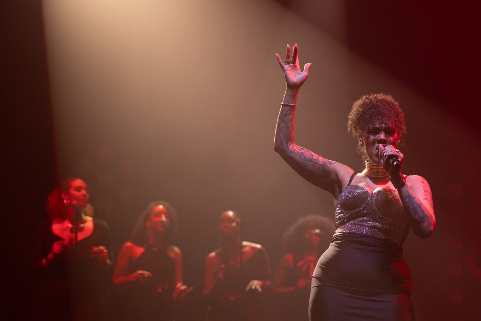A vocalist with her arm raised on a red-lit stage in front of the band