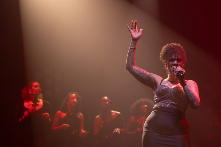 A vocalist with her arm raised on a red-lit stage in front of the band, Les Cigales