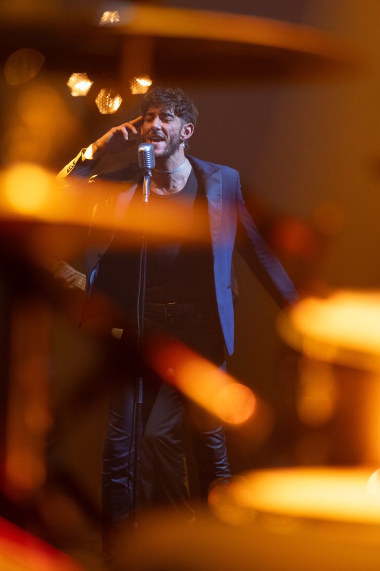 A male singer in a blazer in front of a drum kit in amber light, Les Cigales